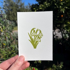 May include: A white card with a green floral design featuring snowdrop flowers. The illustration is a simple, botanical print. The card is held in front of a blurred background of green foliage and a blue sky.