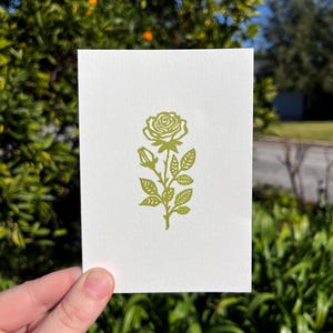 May include: A white card featuring a green rose illustration. The rose is depicted with detailed petals, leaves, and buds. The card is held against a backdrop of green foliage and a bright blue sky.