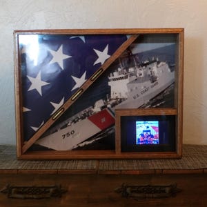 Pode incluir: Uma caixa de sombra de madeira com uma bandeira americana dobrada, uma foto de um navio da Guarda Costeira dos Estados Unidos e uma foto menor de um grupo de pessoas uniformizadas. O navio está etiquetado como "U.S. Coast Guard" e a foto menor está em uma moldura.