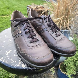 May include: A pair of brown leather sneakers with dark brown laces. The sneakers have a stitched design and the brand name "MEPHISTO" is visible on the side. The shoes are on a black and white surface.