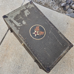 May include: An olive green vintage trunk with a circular emblem featuring a star and wings. The trunk has metal hardware, including corner protectors and clasps. The trunk's surface shows signs of wear and age.