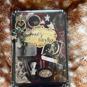 Puede incluir: Una funda protectora transparente para lector electrónico con un collage de imágenes de academia oscura que incluye un busto, una taza de cacao y texto. Un anillo de metal plateado está sujeto en la esquina superior izquierda, adornado con pequeños dijes que incluyen una mariposa, una flor y cuentas verdes. La etiqueta central dice Anne's Kindle.