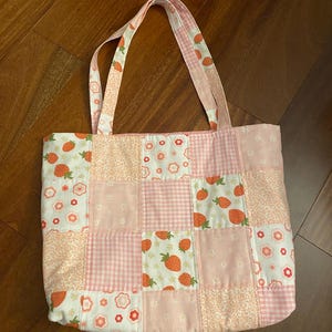 以下が含まれることがあります： イチゴ柄のピンクと白のパッチワークトートバッグ。バッグは2本のストラップが付いており、コットン生地で作られています。