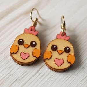 Könnte beinhalten: Ein Paar Ohrringe aus Holz in Form eines Küken mit einem rosa Herzen auf der Brust. Die Ohrringe sind in Gelb, Orange und Rosa bemalt.