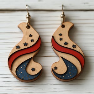 Op de afbeelding: Een paar houten oorbellen met een rood, wit en blauw ontwerp. De oorbellen zijn gevormd als een halve maan met sterren en een rode streep.