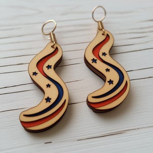 Op de afbeelding: Een paar houten oorbellen met een rood, wit en blauw ontwerp. De oorbellen zijn gevormd als een golvende lijn met uitgesneden sterren. De oorbellen zijn bevestigd aan gouden haken.