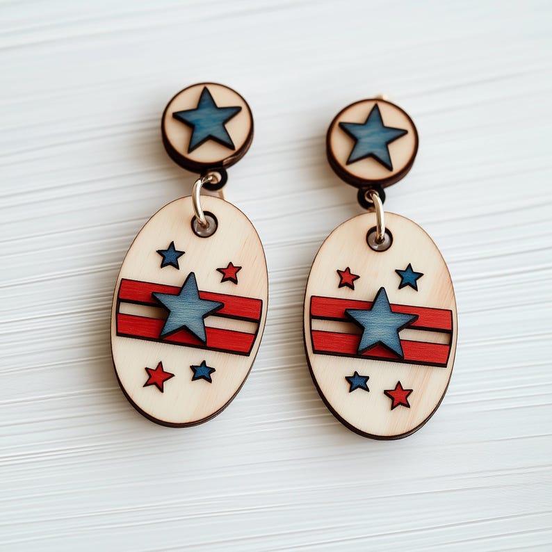 Op de afbeelding: Een paar houten oorbellen met een rood, wit en blauw sterrendesign. De oorbellen zijn ovaalvormig en hebben bovenaan een klein sterretje.