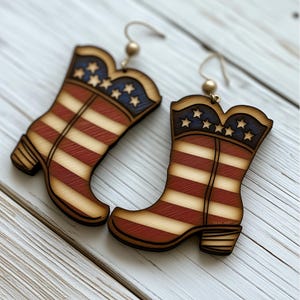 Op de afbeelding: Een paar houten oorbellen in de vorm van cowboylaarzen met een Amerikaans vlagontwerp. De laarzen zijn rood, wit en blauw met witte sterren aan de bovenkant.