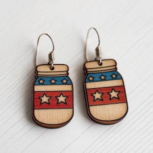Op de afbeelding: Een paar houten oorbellen in de vorm van glazen potten met een rood, wit en blauw Amerikaans vlagontwerp. De oorbellen hebben zilveren haken.
