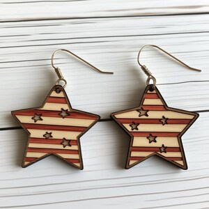 Op de afbeelding: Een paar houten stervormige oorbellen met een rood-wit gestreept patroon en kleine witte sterren. De oorbellen hebben zilveren haakjes.