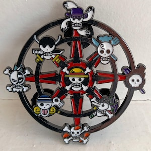 May include: A black metal pin with a compass design featuring eight different skull characters. The characters are all wearing different hats and have different facial expressions. The compass is surrounded by a red and black design.