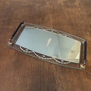 May include: Rectangular glass tray with a mirrored surface and decorative scalloped edges. The tray features black handles with brass accents on each end. A floral design is etched on the mirrored surface.