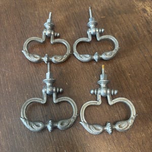 May include: Four antique silver-toned drawer pulls with a decorative, ornate design. Each pull features a curved handle with floral-like detailing and a threaded post for mounting. The pulls are arranged on a wooden surface.