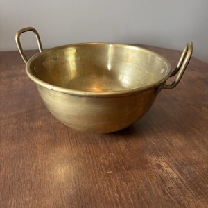 May include: A round, brass bowl with two curved handles. The bowl has a slightly aged appearance, with a warm, golden hue. The handles are attached to the sides of the bowl, providing a secure grip. The bowl is sitting on a wooden surface.