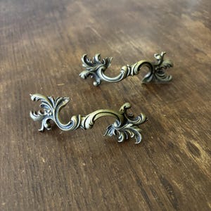 May include: Two antique-style drawer pulls with an ornate, floral design. The pulls are made of a brass-colored metal and have a curved handle shape. The background is a dark wooden surface.