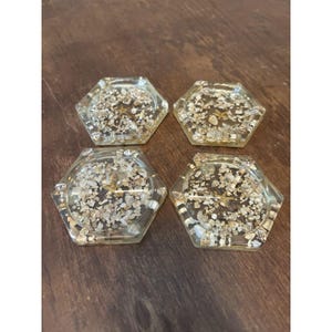 May include: A set of four hexagonal coasters made of clear resin, each containing embedded small white and tan seashells and a central starfish. The coasters have a smooth, glossy finish and a transparent appearance, showcasing the natural textures of the shells.