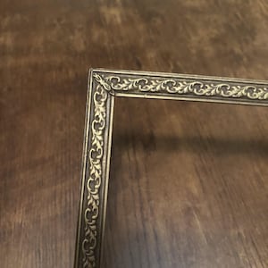 May include: An antique-style metal picture frame corner featuring a raised, intricate leaf and vine relief pattern in a muted gold tone. The frame has a rectangular profile with a mitered corner joint, showcasing a detailed, embossed texture.