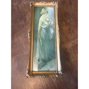 May include: A framed print featuring a woman in a long dress holding a baby. The artwork is in shades of green and gold, with the woman wearing a yellow shawl. The ornate gold frame adds a vintage touch to the piece.