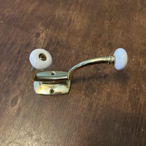 May include: A dual-pronged wall hook featuring a polished gold-tone metal base and curved arm. The hook is accented with two smooth, round white ceramic knobs at the ends of the prongs. The mounting plate includes two pre-drilled screw holes.