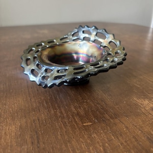 May include: A small, iridescent glass bowl with a decorative, openwork rim. The bowl's interior has a metallic sheen with colors of purple, gold, and green. The rim is silver and black, creating a contrast. The bowl sits on a wooden surface.