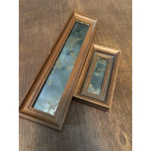 May include: Two rectangular wall mirrors with medium-toned wooden frames. The glass surfaces feature a dark, cloudy gray background with an intricate, weathered gold-colored leaf or vine pattern overlaying the reflective surface.