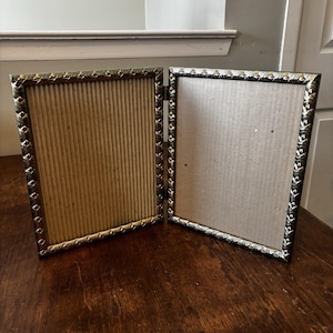 May include: A hinged, gold-toned metal picture frame, open to display two rectangular openings. The frame has a decorative, textured border. The backing is a light brown, corrugated cardboard. The frame is on a dark wooden surface.
