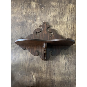 May include: A dark brown, antique-style wooden wall shelf. The shelf has a curved front and decorative, carved details. It features a small shelf supported by a central bracket, perfect for displaying small items. The wood grain is visible.