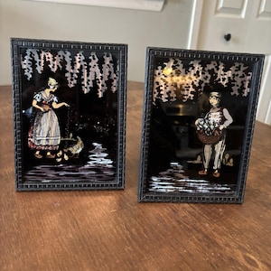 May include: Two rectangular black-framed wall art pieces featuring reverse-painted glass scenes. The left piece shows a person in a striped skirt feeding chicks. The right piece depicts a person holding a basket of flowers against a dark background.