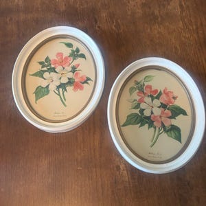 May include: Two vintage framed floral prints. Each print features a bouquet of pink and white flowers with green leaves, set against a beige background. The oval frames are white with a gold inner border.