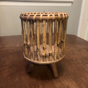 May include: A cylindrical bamboo plant stand featuring vertical bamboo stalks arranged in a cage-like structure, a flat circular base, and three short, sturdy wooden legs. The natural light brown bamboo has visible nodes and a textured finish.