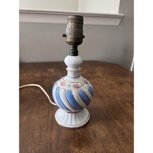 May include: A small, vintage table lamp with a white base and a blue and white striped globe. The globe is decorated with pink floral accents and gold trim. The lamp has a brass bulb socket and a white cord.