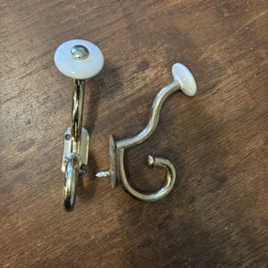 May include: Two wall hooks made of polished gold-tone metal, each featuring a smooth white ceramic knob at the top. The hooks have a curved design with a mounting plate and a visible screw, providing a functional structure for hanging items.