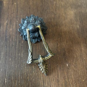 May include: Antique brass drawer pull with a floral backplate and a decorative handle. The handle has a leaf design. The hardware is set against a wooden surface.