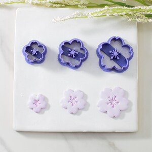 May include: Three purple plastic flower-shaped cookie cutters and three purple fondant flower shapes on a white surface.