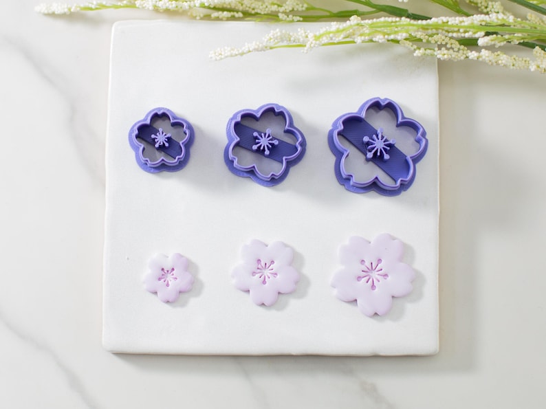 May include: Three purple plastic flower-shaped cookie cutters and three purple fondant flower shapes on a white surface.