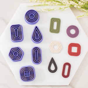 May include: A collection of polymer clay cutters in various geometric shapes, including circles, rectangles, and teardrops. The cutters are in shades of purple, green, red, and beige, arranged on a white hexagonal surface. Decorative floral accents are in the background.