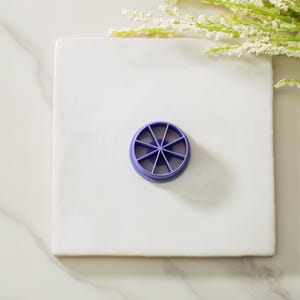 May include: Purple plastic round cookie cutter with eight points, on a white surface.