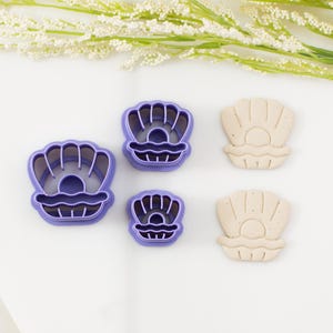 May include: Two purple plastic shell-shaped cookie cutters and two corresponding beige clay cutouts. Each piece features a clam shell design with a central pearl. The cutters have raised edges for pressing into dough or clay to create detailed impressions.