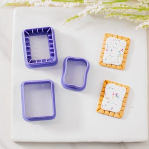May include: A collection of purple plastic cookie cutters in various rectangular shapes, alongside two miniature pastry replicas with white frosting and colorful sprinkles. The items are arranged on a white surface.