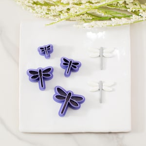 May include: A collection of dragonfly-shaped cookie cutters in various sizes. The cutters have a purple outline with a black interior, and two white dragonfly decorations are also present. The items are arranged on a white square surface.