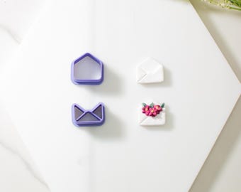 Può includere: Due tagliapasta a forma di busta in plastica viola e due buste in pasta di zucchero bianca, una semplice e l'altra decorata con fiori rosa.