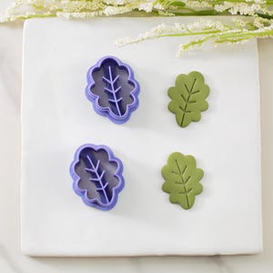 May include: Two leaf-shaped cookie cutters, one in purple and the other in green, sit on a white surface. The purple cutters have a detailed leaf vein design. Two green leaves are also present, showcasing the same leaf vein design.