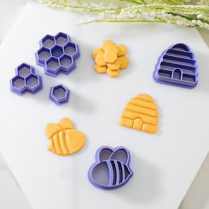May include: A set of purple plastic cookie cutters in the shape of a bee, honeycomb, and beehive. There are also two yellow sugar cookies, one shaped like a bee and the other like a beehive.