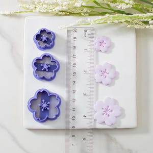 May include: Set of three purple plastic flower cookie cutters with three matching purple fondant flowers of different sizes. The cutters are 1.5 inches, 2 inches, and 2.5 inches in diameter.