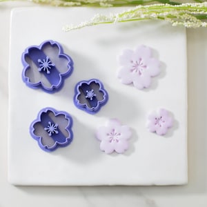May include: Five purple plastic flower-shaped cookie cutters and three purple fondant flowers on a white surface.