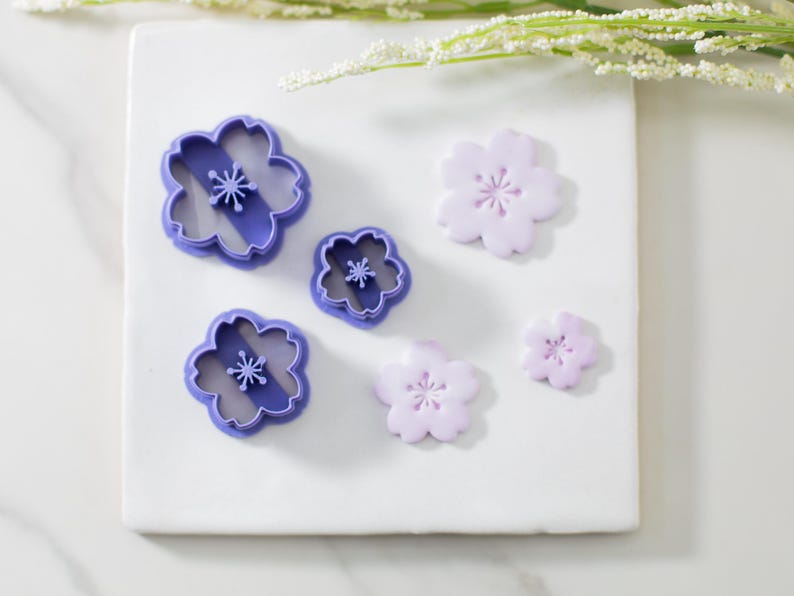 May include: Five purple plastic flower-shaped cookie cutters and three purple fondant flowers on a white surface.