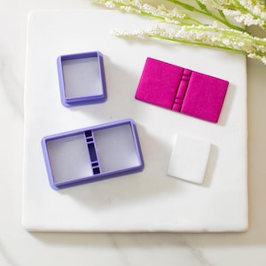 May include: Three purple plastic cookie cutters on a white surface. The cutters are shaped like a book, a square, and a rectangle with a line down the middle.