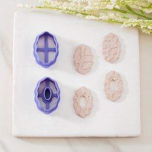 May include: An overhead shot displays two purple polymer clay cutters and four beige clay earrings. The cutters have an oval shape with interior designs. The earrings have textured surfaces and various oval shapes. White flowers are in the background.