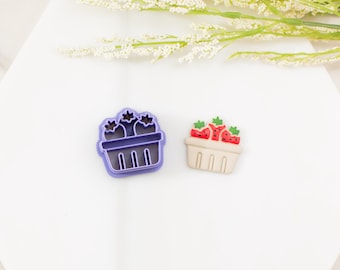 Può includere: Un tagliabiscotti in plastica viola a forma di cestino di fragole accanto a un pezzo finito in argilla o resina che raffigura un cestino beige riempito con tre fragole rosse con foglie verdi e semi neri su una superficie bianca.
