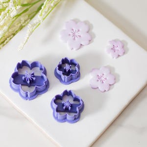 May include: Five purple plastic flower-shaped cookie cutters of varying sizes, with three pink fondant flowers on a white surface.